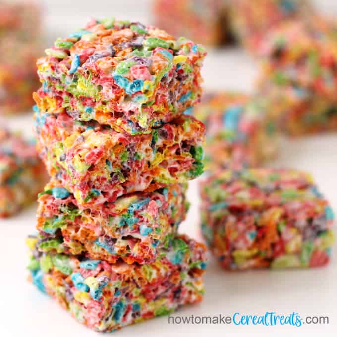A close-up image of squares of Fruity Pebbles rice crispy treats stacked up and scattered around on a white table. 