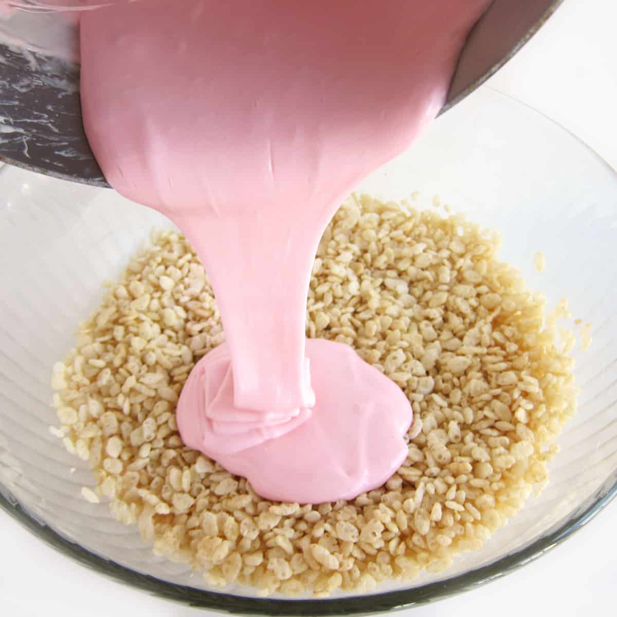 pouring light pink-colored melted marshmallows over a bowl of Rice Krispies