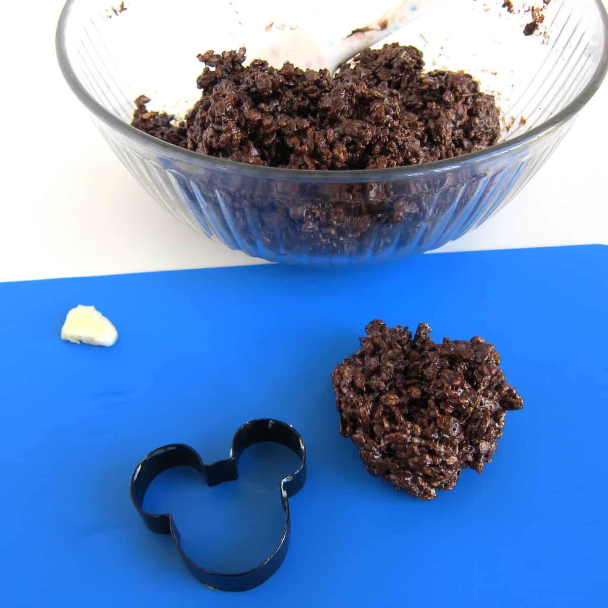 Mickey Mouse cookie cutter greased with butter next to chocolate rice krispie treat mixture