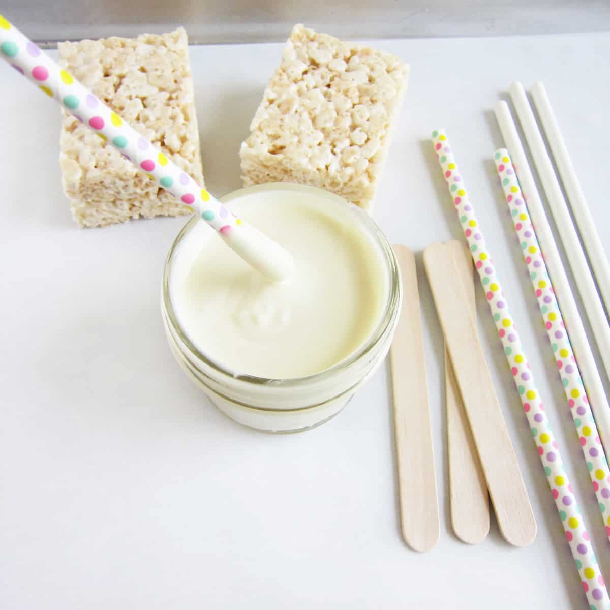 Dipping a polka dot paper straw into a bowl of white chocolate on a tray with rice krispie treats, popsicle sticks, and lollipop sticks.