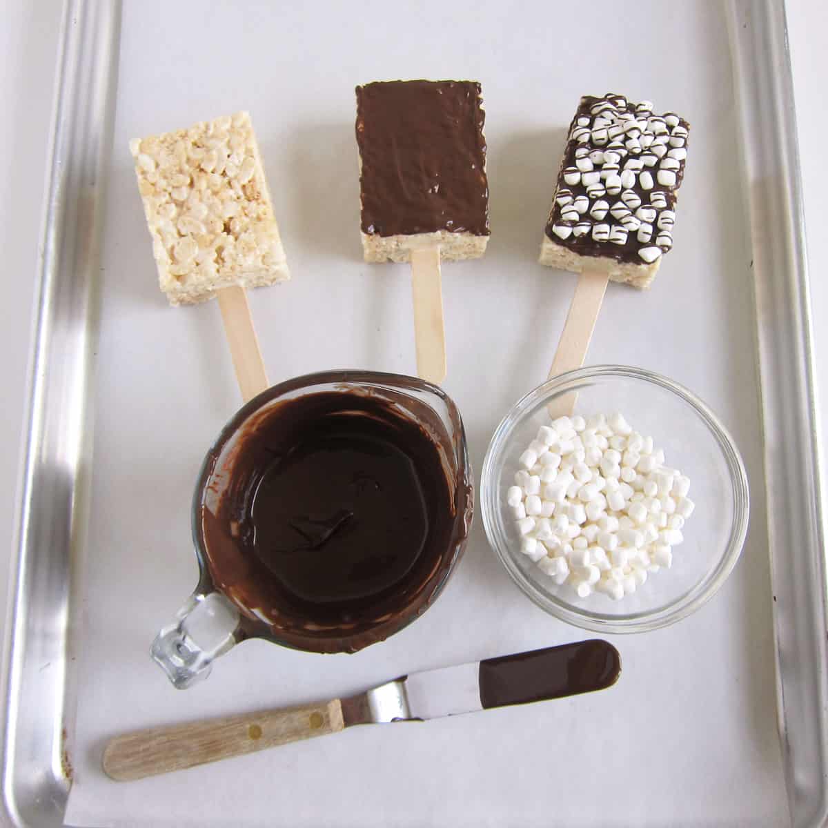 Rice Krispie Treat pops plain, covered in chocolate, and topped with dehydrated marshmallows next to bowls of melted chocolate and tiny marshmallows.