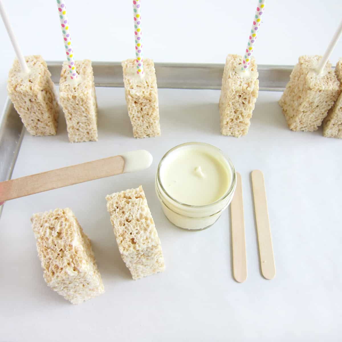 Popsicle stick dipped in melted white chocolate on a tray with Rice Krispie Treat pops.