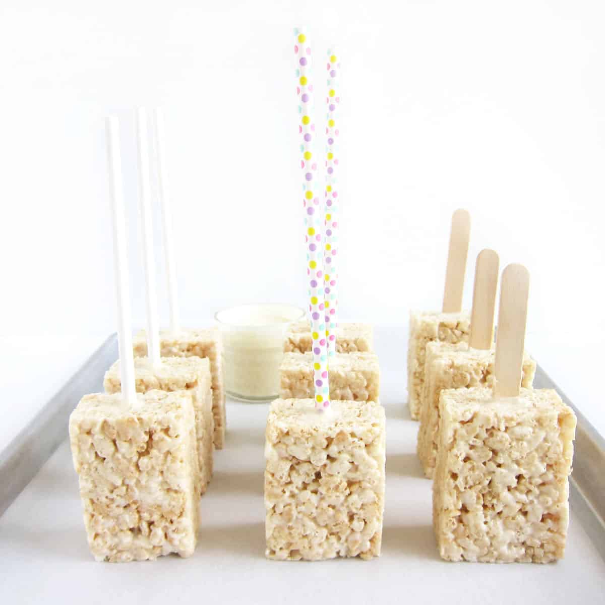 A variety of Rice Krispie Treat pop with lollipop sticks, paper straws, and popsicle sticks on a baking tray.