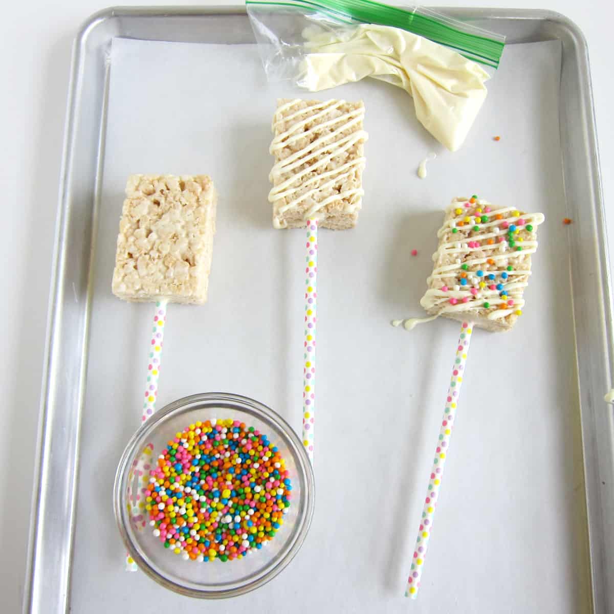 Plain rice krispie treat, one drizzled with white chocolate, and one topped with rainbow Crispearls sprinkles next to a bowl of Crispearls and a zip-top bag of melted white chocolate.