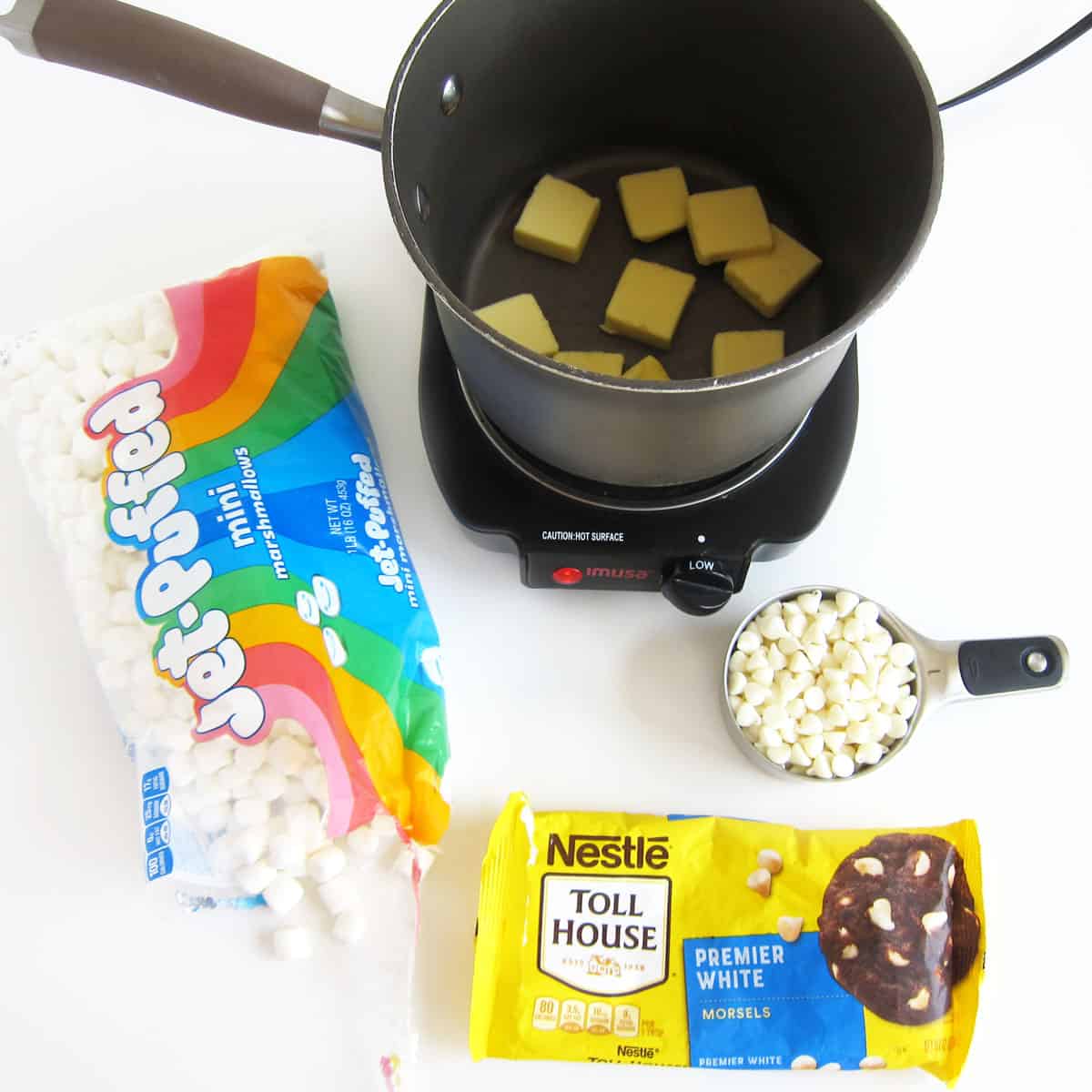 Slices of butter in a saucepan set over low heat on a stovetop alongside a bag of Jet-Puffed Mini Marshmallows and Nestle Premier White Morsels.