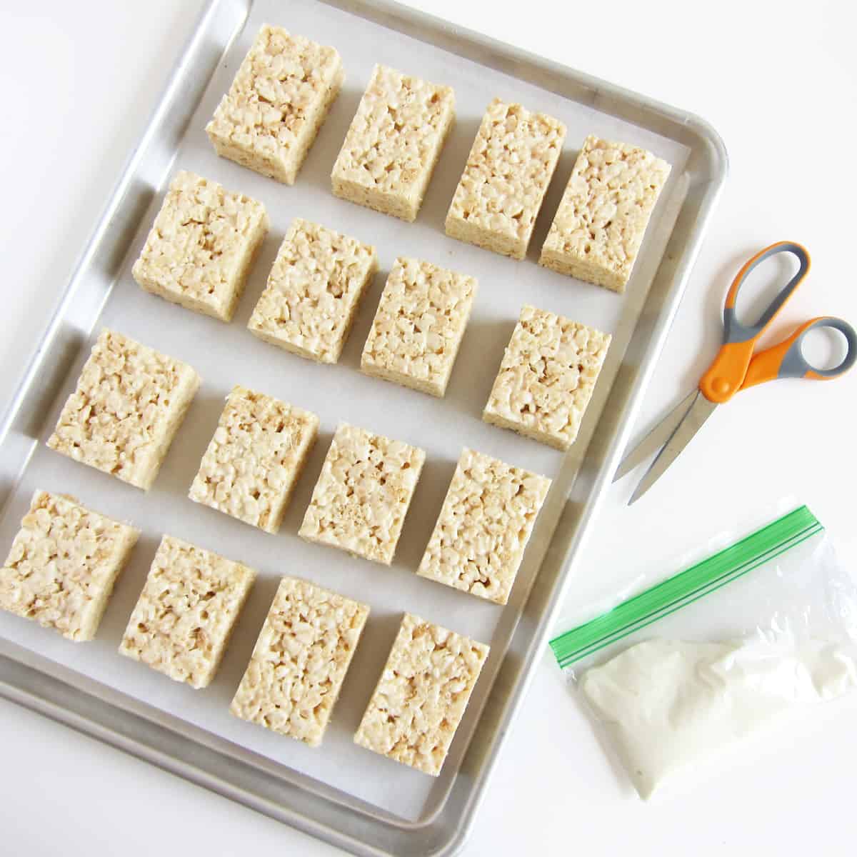 white chocolate rice crispy treats on a tray next to a pair of scissors and a zip-top bag of melted white chocolate