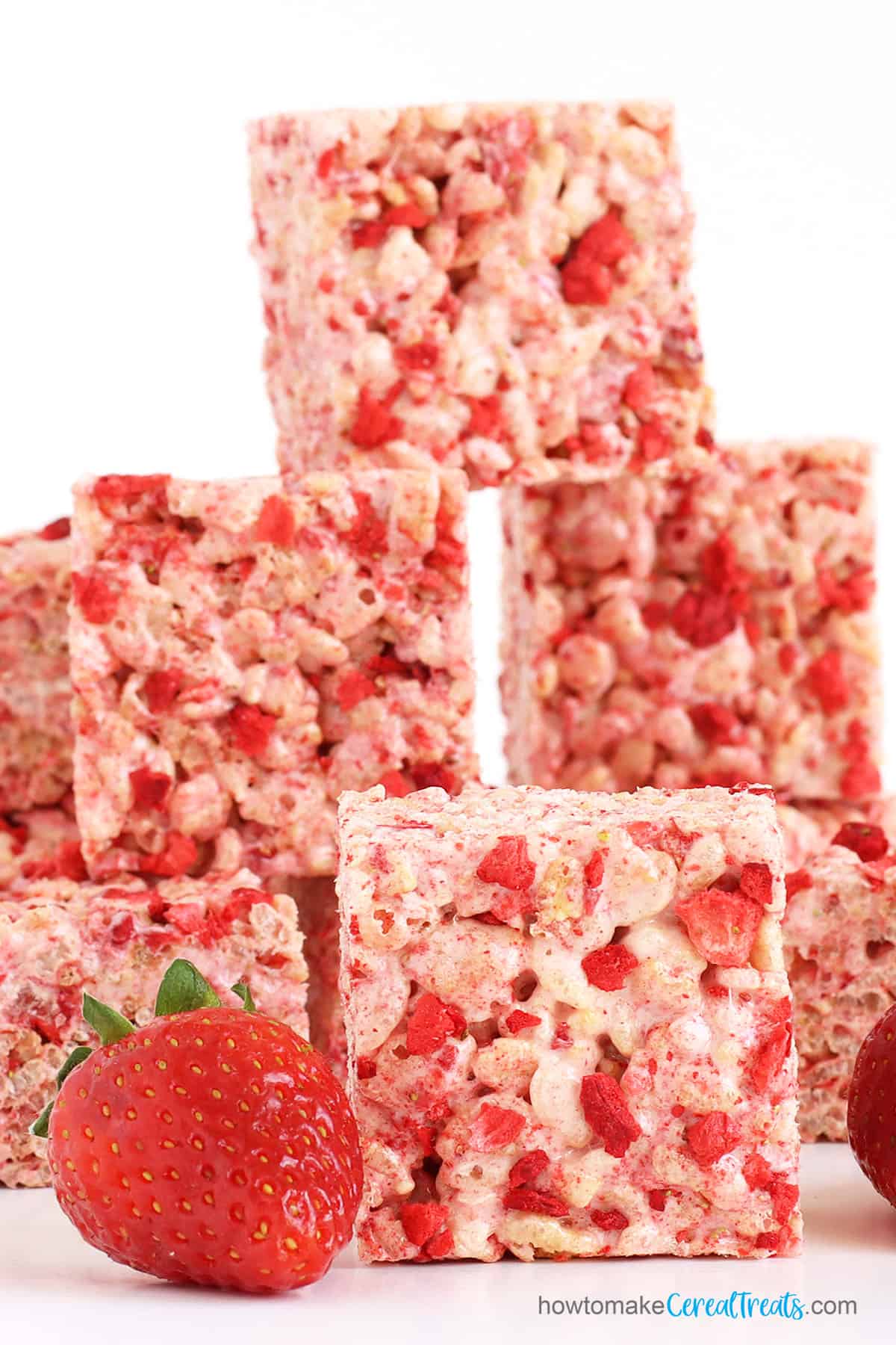 Square rice krispie treats packed with freeze-dried strawberries are stacked up next to fresh strawberries.