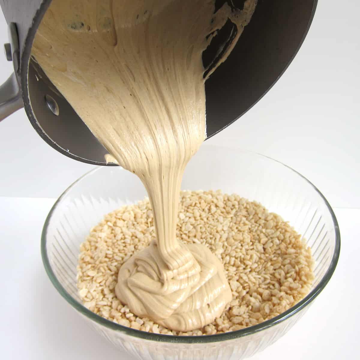 pouring the mixture of peanut butter, corn syrup, and marshmallow fluff over a bowl of Rice Krispie Treats cereal.