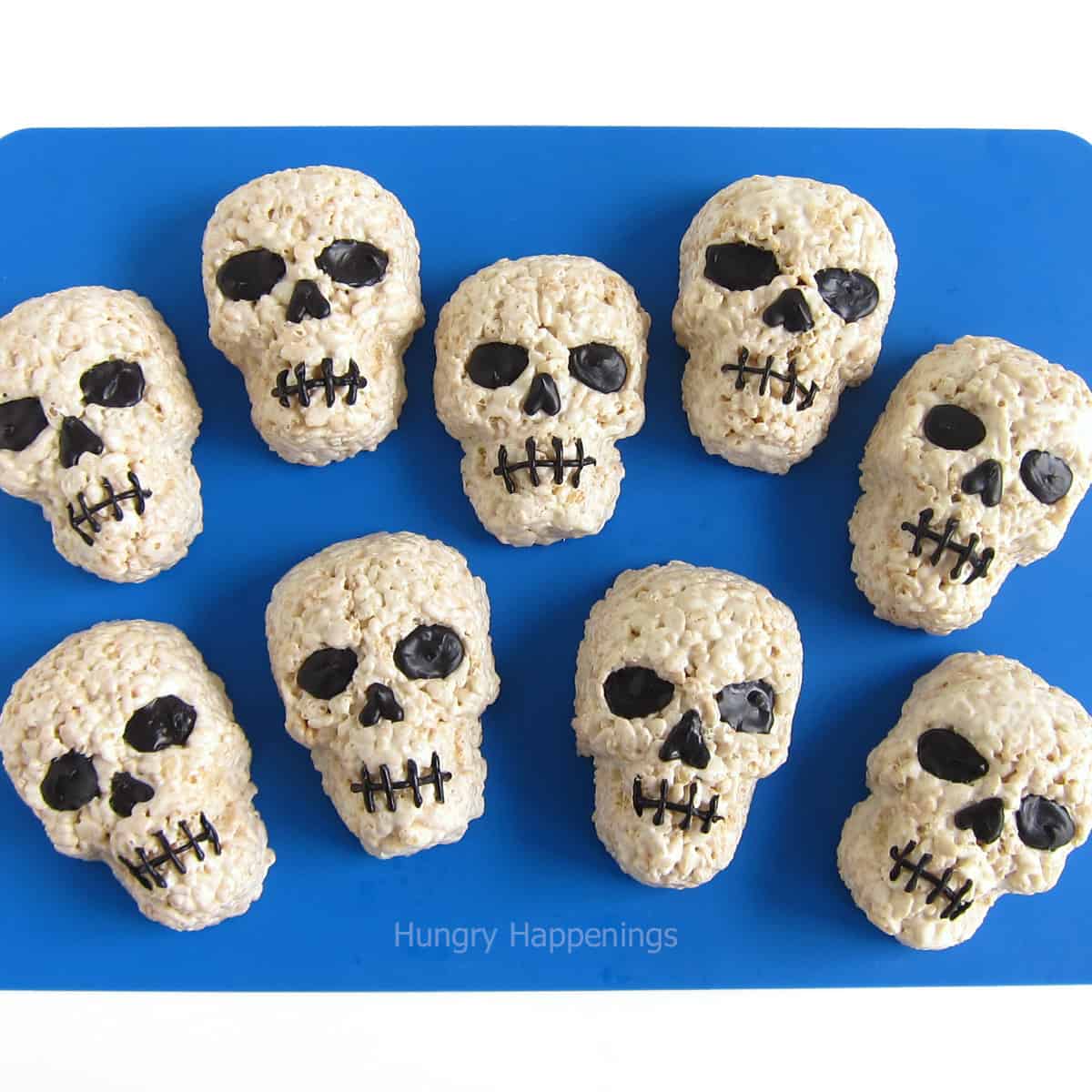 white chocolate rice krispe treat skulls decorated with black eyes, nose, and mouth set on a blue silicone mat.