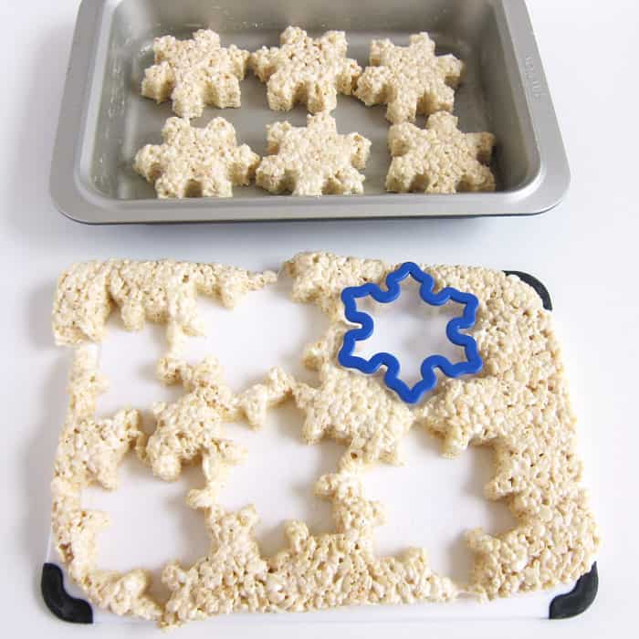snowflake rice krispie treats in pan next to a cutting board with cut-out rice krispie treat snowflakes.